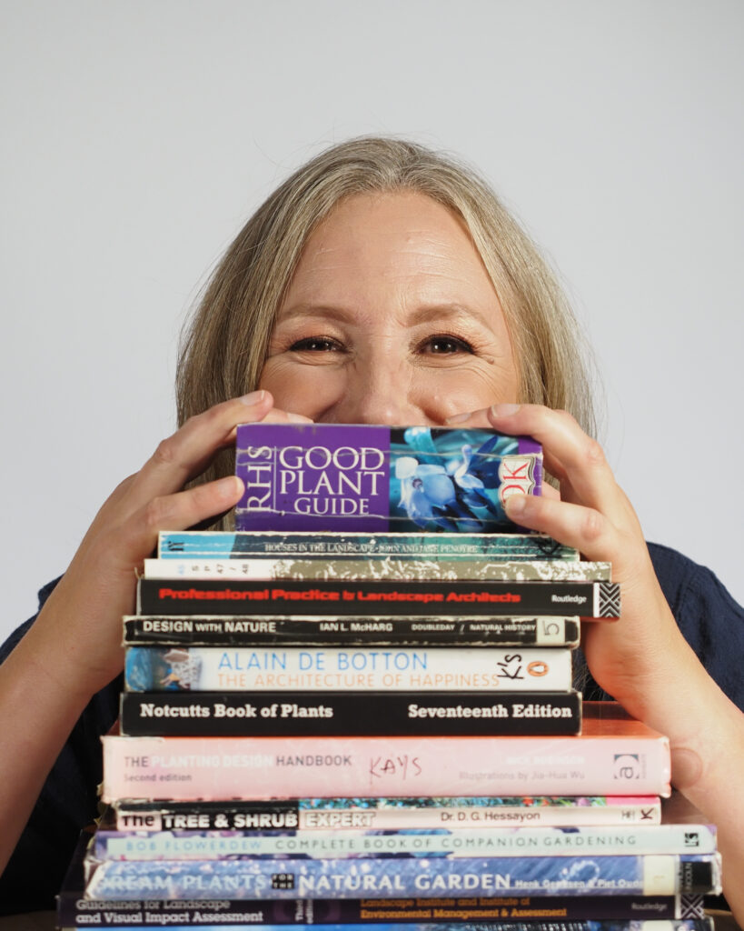 Kay. Portrait of a woman peering over a pile of books. She's smiling. White background.